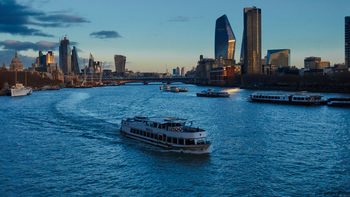Thames skyscrapers ferry This urban landscape photograph was taken in the evening during winter and shows a section of the River Thames in London, United Kingdom. The main subject of the image is a ferry named "Mercia" navigating the river, surrounded by several other boats. Prominently featured in the background are notable examples of London architecture, including the Gherkin (30 St Mary Axe) and Cheese Grater (122 Leadenhall Street), contributing to the distinctive skyline. The scene is further enhanced by modern and historic buildings along the riverbank, with light reflecting off the water and skyscrapers, illustrating the blend of contemporary and historical elements characteristic of London.